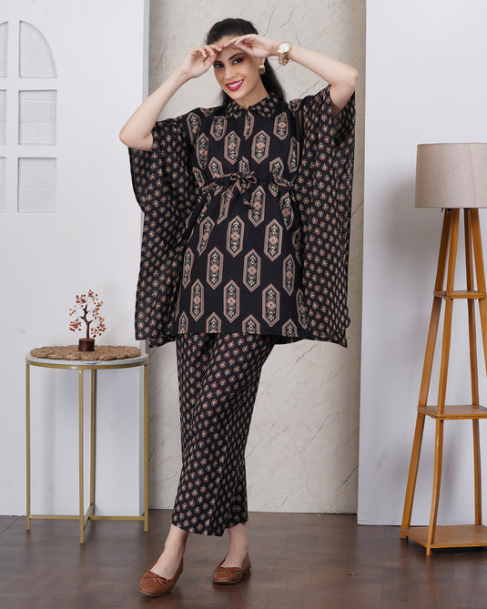 A woman in black two-piece kurta set with beige and gold motifs,standing in a styled indoor room with a side table, lamp, and marble wall panel.