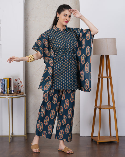 A woman models a dark teal blue three-piece kurta set with a geometric pattern, featuring a tunic top, matching pants, and a dupatta, styled in an indoor setting with a lamp and side table.