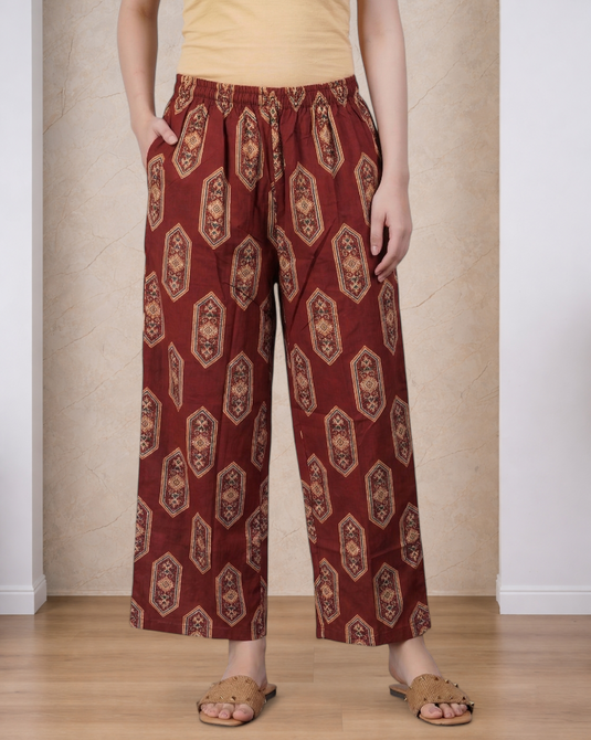 A person wearing loose, wide-leg cropped pants in a rust red color with a beige and brown geometric pattern, paired with a beige top and brown slide sandals, standing against a beige textured wall and light wood floor.