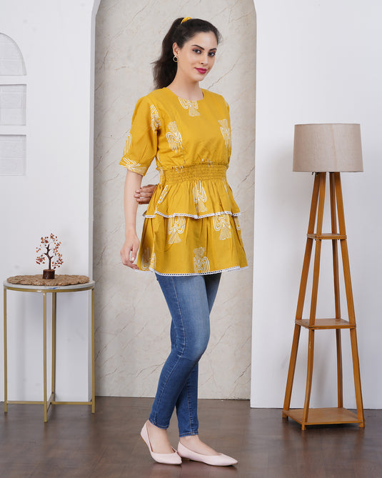 Woman wearing a yellow top and blue jeans standing in a room with a aesthetic lamp and small table.