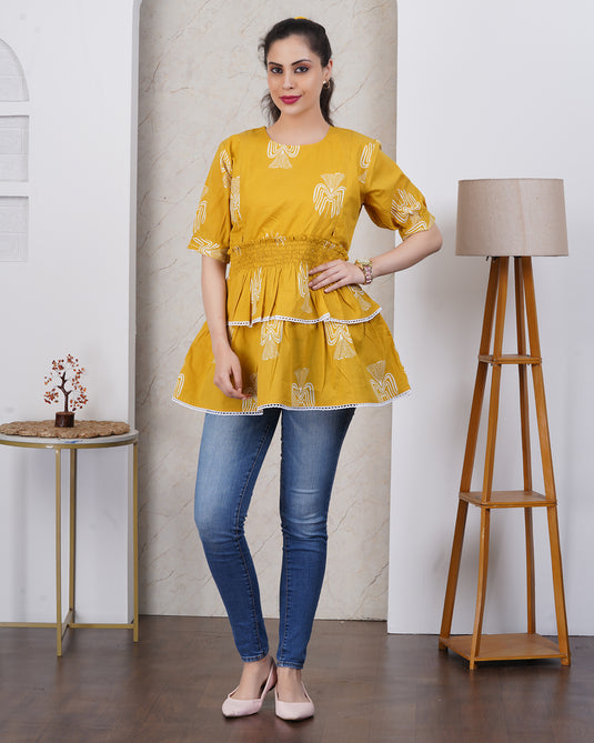 Woman wearing a yellow top with white patterns and blue jeans standing in a room with a lamp and small table.