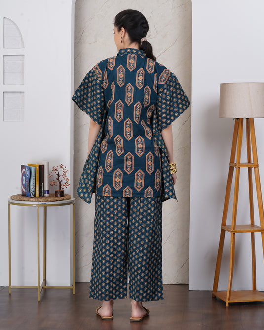 A woman stands with her back to the camera wearing a dark teal two-piece outfit with a beige and orange geometric pattern. The set includes a short-sleeved button-up shirt and matching wide-leg pants.