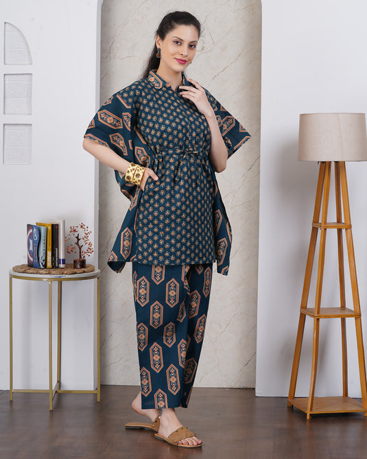 A woman models a dark teal blue and gold printed two-piece kurta set with pocket in a styled indoor room with a marble column, side table, and floor lamp.