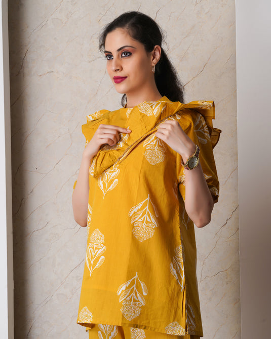 Woman wearing a yellow feeding outfit with two nursing zips and white patterns against a beige wall.