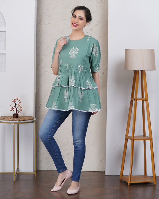 Pregnant Woman wearing a green and white motif maternity top and blue jeans standing in a room with a lamp and small table.