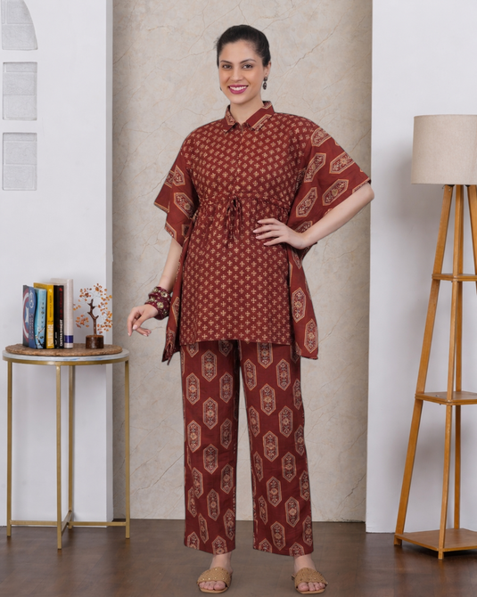 A woman models a two-piece rust-colored printed kurta set in a styled indoor setting. The set features a short-sleeve, collared kurta with a drawstring waist and matching wide-leg trousers, both decorated with a geometric and paisley pattern.