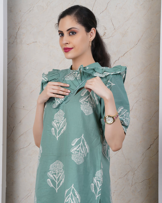 Woman wearing a green nursing dress with two feeding zippers in floral patterns against a beige wall.