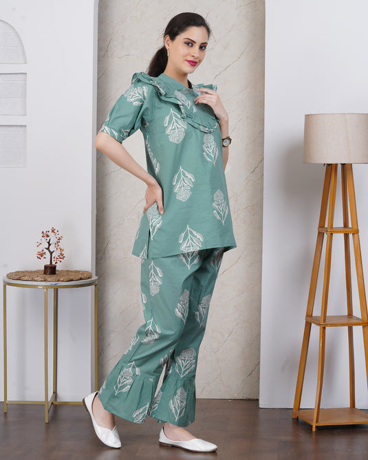 Woman wearing a green patterned outfit in a room with a lamp and small table.