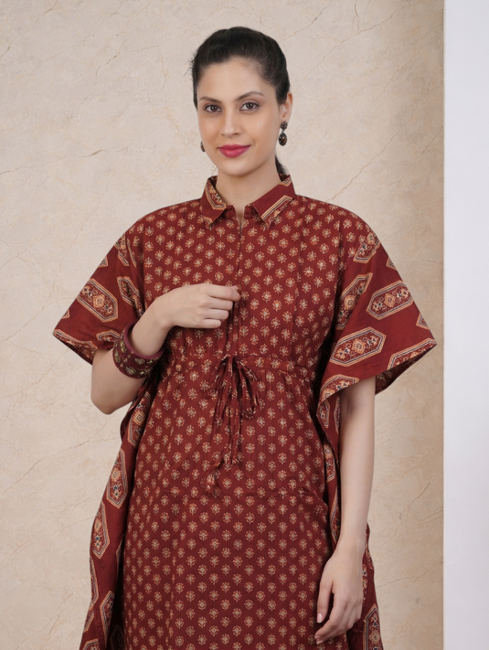 A woman modeling a rust-red printed kurta with a tie waist and short sleeves, set against a textured beige background.