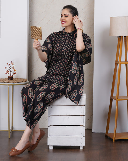 A woman sits on a white wooden crate in a styled indoor setting, wearing a black printed kurta and matching pant set with beige geometric motifs. She holds a small mirror in her right hand, with a floor lamp, side table, and marble wall paneling in the background.