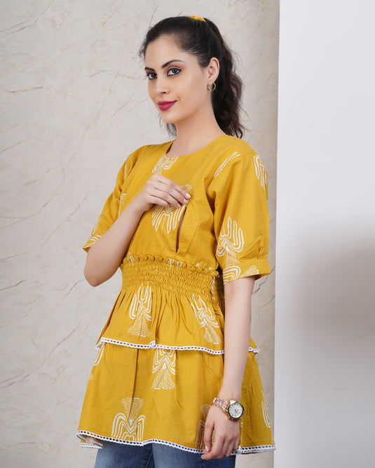 Woman wearing a yellow embroidered nursing top with two feeding zips against a light gray wall.