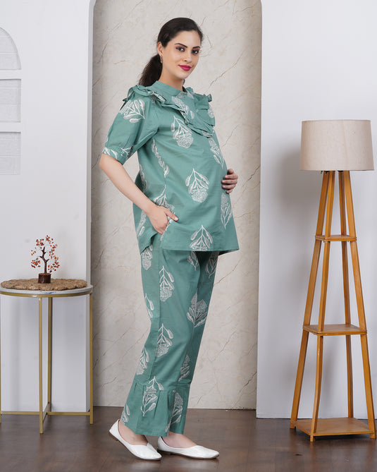 Pregnant Woman wearing a green floral maternity set standing in a room with a lamp and small table.