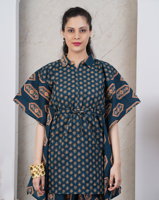 A woman modeling a dark teal blue printed tunic dress with beige and orange geometric patterns, featuring a collar, button-down front, and wide short sleeves, set against a light beige marble wall.