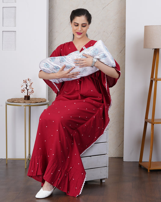 Woman in a red maternity kaftan holding a baby in a room with a white wall and wooden furniture.