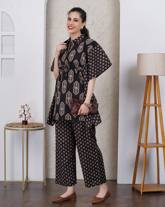 A woman models a black two-piece kurta set with beige and gold geometric patterns, consisting of a short-sleeve tunic top and matching wide-leg trousers, styled with a dark purple clutch and brown flats in an indoor setting.