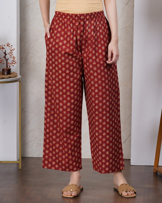 A person wearing loose, wide-leg cropped pants in a rust red base with a repeating small floral pattern in yellow and white. The pants are styled with a beige top and brown slide sandals, set against an indoor background with a marble wall and wooden floor.