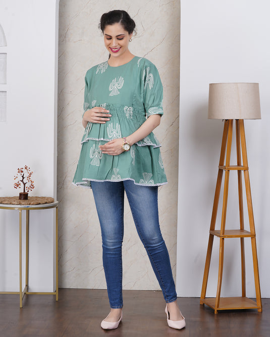 Pregnant Woman with bump wearing a green maternity top and blue jeans standing in an aesthetic room with a lamp.