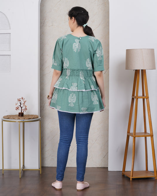 Woman wearing a green tiered dress with white patterns in a room with a lamp and small table.
