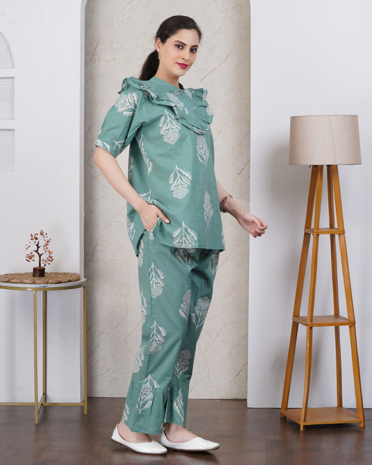 Woman wearing a green floral outfit with pocket standing in a room with a lamp and small table.