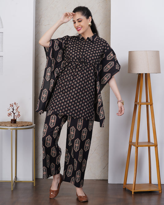 A woman models a black three-piece kurta set with beige and brown geometric patterns, featuring a short-sleeve tunic, matching pants set against an indoor background with a floor lamp and side table.