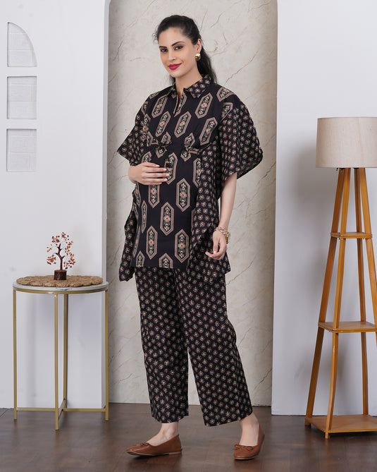 A woman models a black two-piece kurta set with beige and brown geometric patterns, standing in a styled indoor setting with a side table, lamp, and marble wall panel.