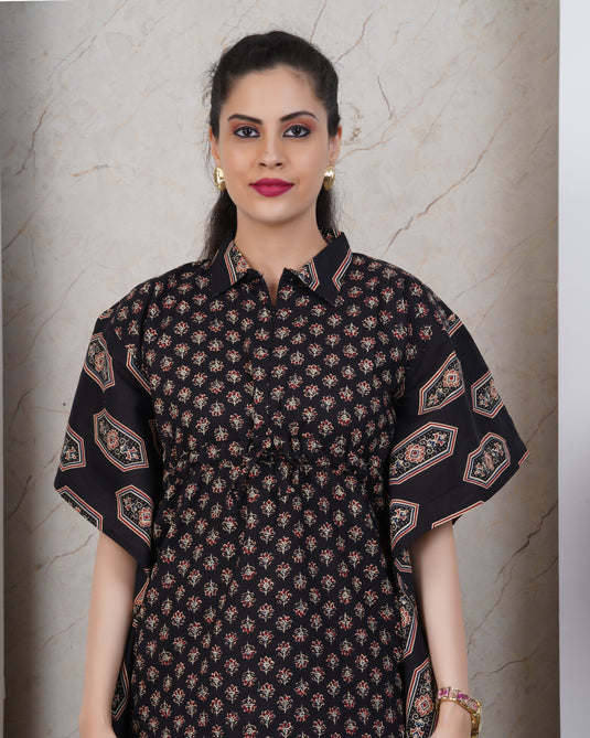 A woman modeling a short-sleeve button-up kurta with a black base and intricate multicolor block print patterns, set against a neutral marble-textured wall.