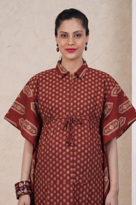 A woman modeling a rust-red printed kurta with a geometric pattern, featuring a pointed collar, button front, drawstring waist, and wide short sleeves with patterned cuffs, set against a beige marble-textured background.